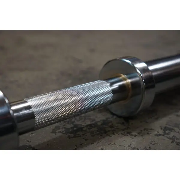 Close-up of a Power Maxx Big Olympic Dumbbell Handle, featuring a textured silver grip and smooth weight collars, resting on a rough floor.