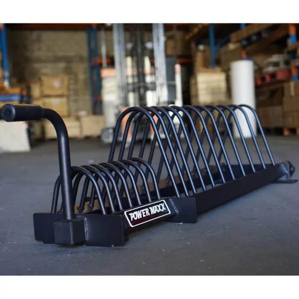 In a warehouse, a Bumper Weight Plate Toaster Rack sits on the concrete floor. Its black metal frame, akin to a sturdy toaster rack, offers slots for organizing weight plates against shelves and boxes.