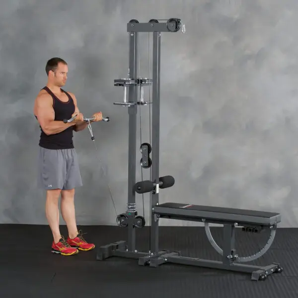 A person in a black tank top and gray shorts is using the Ironmaster Cable Tower Attachment V2 for exercise at home, performing a cable curl with an attached bench.