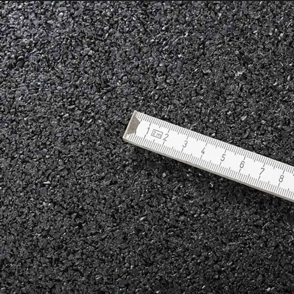 A close-up image of the GymFloor® 15mm 1m x 1m Black surface with a metallic ruler placed diagonally. The ruler, marked in centimeters and partially visible, accentuates the texture and granularity similar to asphalt.