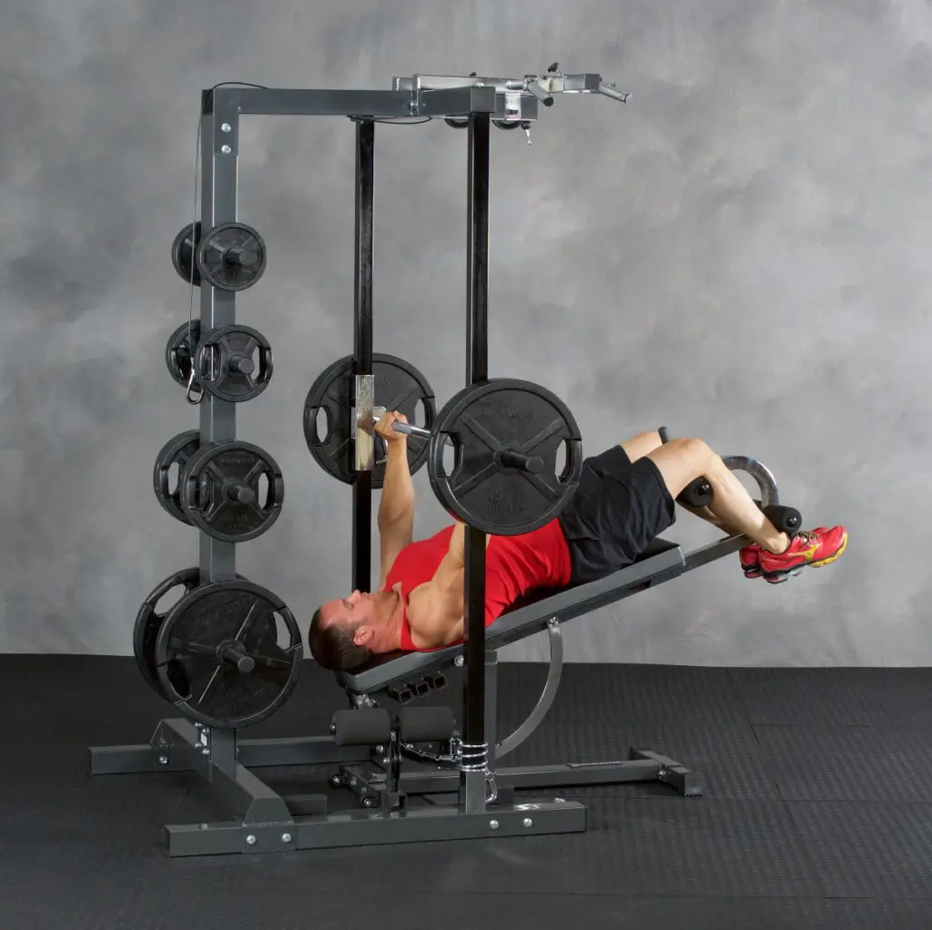 A person in a red tank top and black shorts performs a bench press on a Smith machine, lying on a decline bench with weighted plates on the barbell and rack.