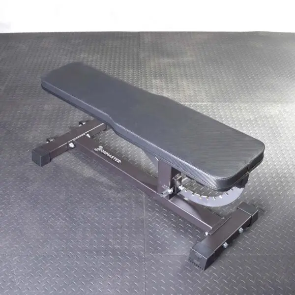 The Ironmaster Hybrid Bench Pad, a black adjustable workout bench with a cushioned pad, rests on a textured gym floor. It is set in a flat position, showcasing visible branding on its sturdy frame.