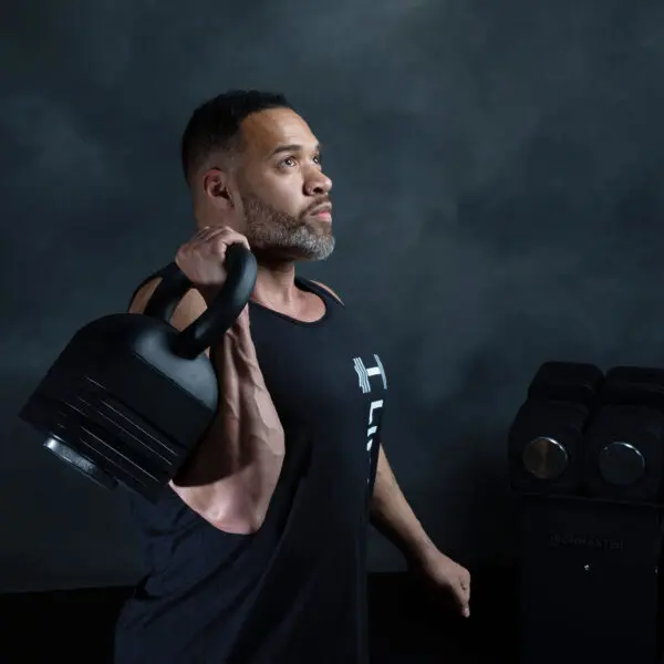 A man in a black tank top holds an Ironmaster Adjustable Kettlebell Handle over his shoulder, looking focused. Behind him, a stack of weights is set against a dark, textured wall.