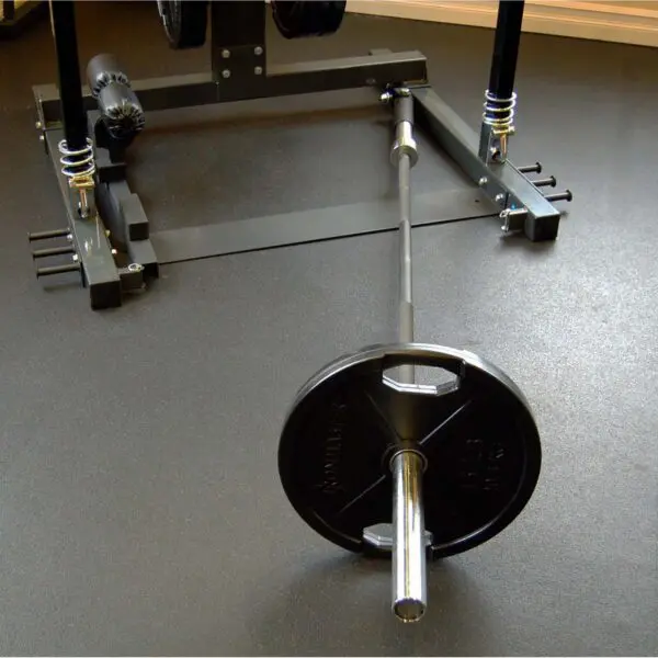 An Ironmaster Landmine Attachment, accompanied by a barbell and weight plates marked 45 lbs, rests on a gyms dark rubberized floor in front of a squat rack, indicating a designated weightlifting area.