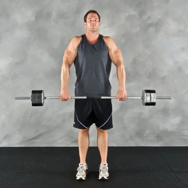 A man in a dark sleeveless shirt and black shorts uses an Ironmaster Straight Barbell to perform an upright row on the gyms black floor against a textured gray background.