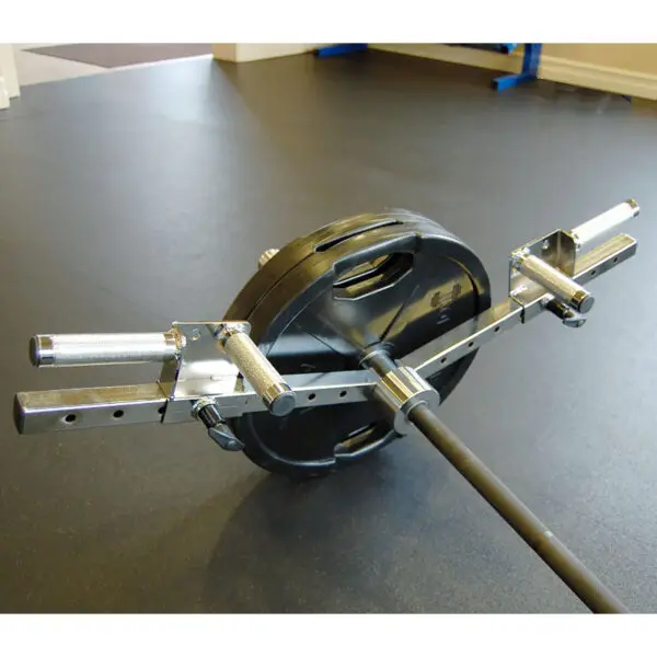 A gym setup features a loaded trap barbell with black weight plates on a rubber gym floor. The Ironmaster Ultimate Row Handle, in chrome, is ready for deadlifts or shrugs. Gym equipment and part of a doorway are visible in the background.