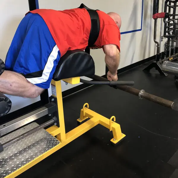 Wearing a red shirt and blue shorts, a person is performing back exercises on a hyperextension bench using the Spud Inc Dr Squat Strap in a gym setting.