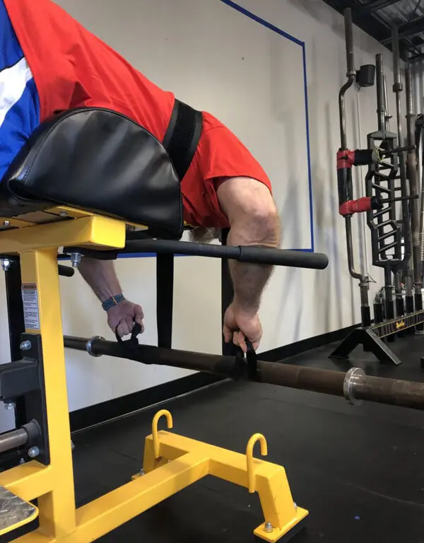 A person in a red shirt is using a Spud Inc Dr Squat Strap in the gym. They lie face down, holding a barbell with their right hand, bending at the waist. Various weights and equipment are visible in the background.