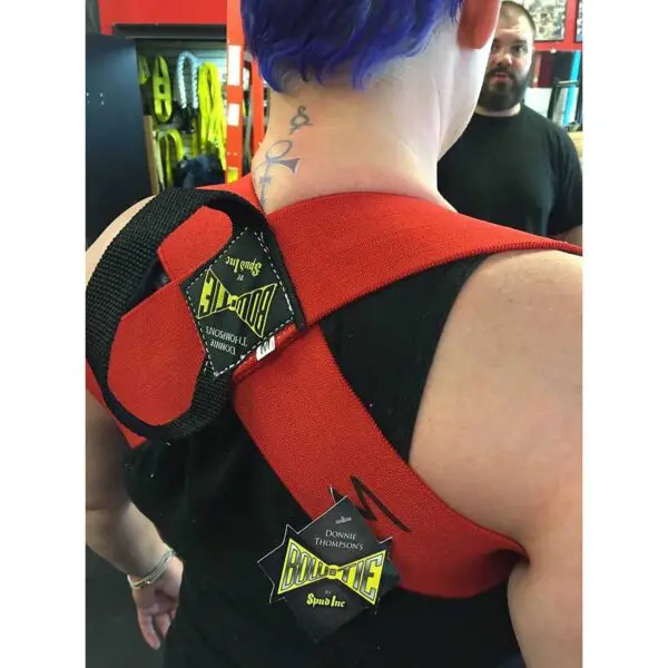A person with purple hair is wearing a red and black Spud Inc Bowtie Casual in a gym setting, with a bearded person in the background.