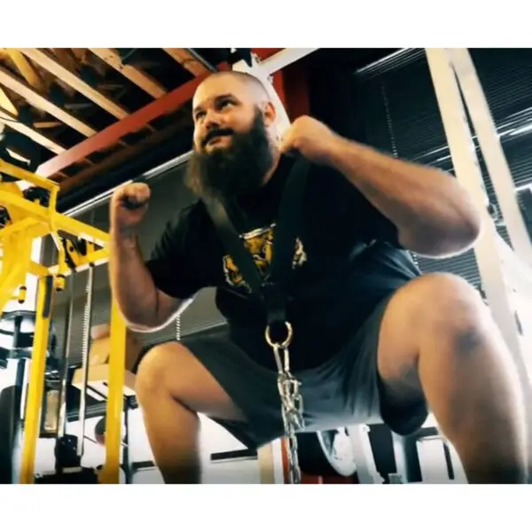 A bearded man is using a Spud Inc. Front Squat Harness for squats in the gym, with equipment and ceiling beams visible in the background.