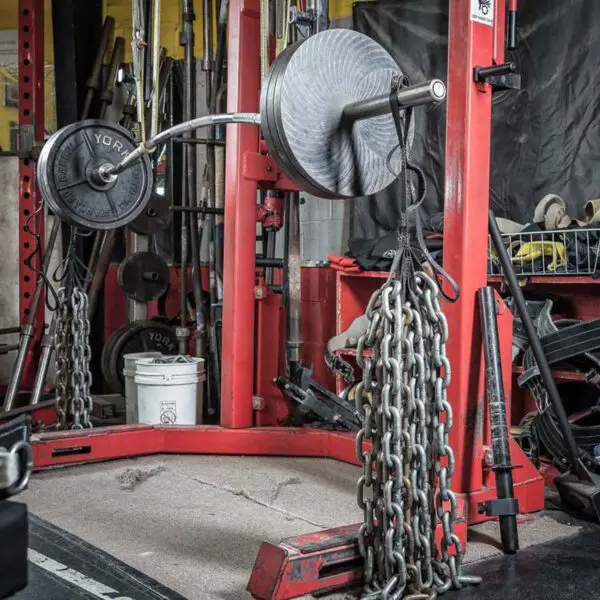 A weightlifting area features a red squat rack with a barbell loaded with weights and chains on each end. Various gym equipment, including Spud Looper Straps, fills the area on the concrete floor.