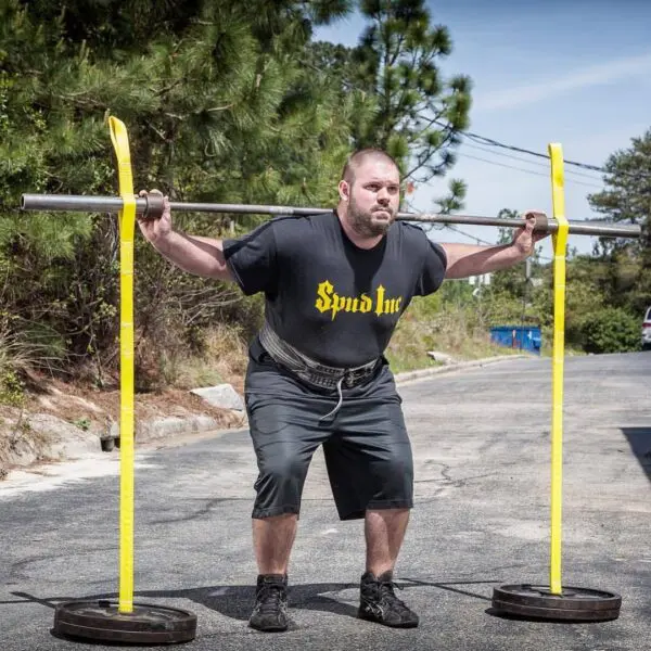 A man is squatting outdoors with a barbell secured by the Spud Inc Swing Set Straps. Hes wearing a black Spud Inc T-shirt, shorts, and sneakers. Trees and a blue dumpster can be seen in the background.