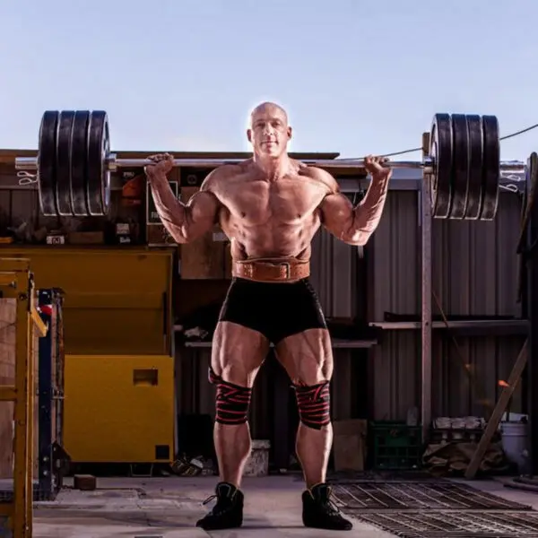 A muscular man performs a heavy barbell squat outside, wearing shorts and the Sting Eco 4 Leather Belt. He has knee wraps and a determined expression, surrounded by gym equipment and a metal structure, all under a clear sky.
