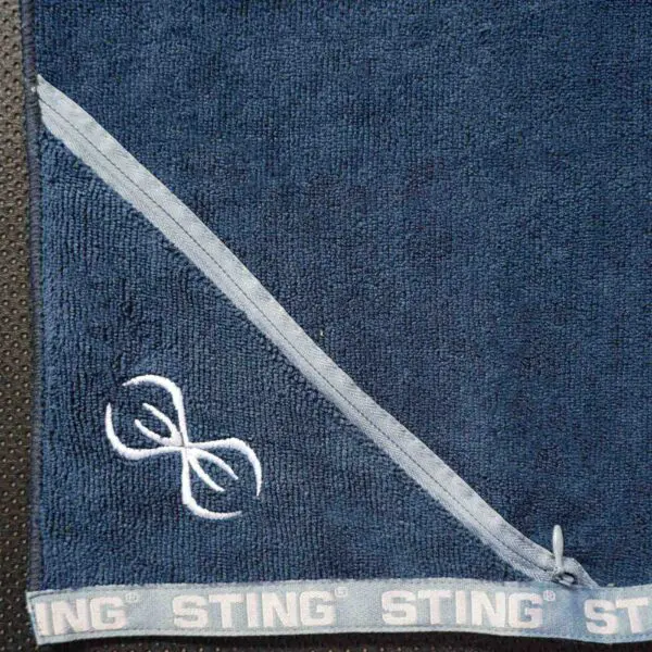 A close-up of the Sting Microfibre Gym Towel in dark blue, featuring a diagonal light blue stripe and a white embroidered logo with two stylized curved lines. The bottom edge repeats the word STING along the border.