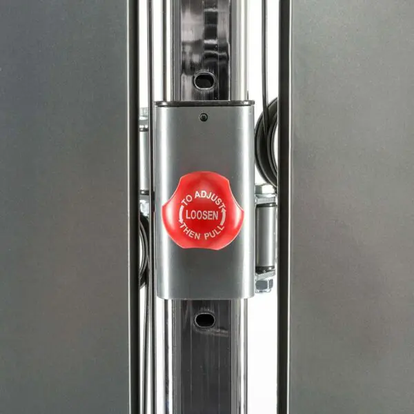 Close-up of a metal adjustment mechanism with a red knob labeled To Adjust Loosen Then Pull. The mechanism is part of a polished metal frame, possibly from a wall mounted cable machine or similar exercise equipment.