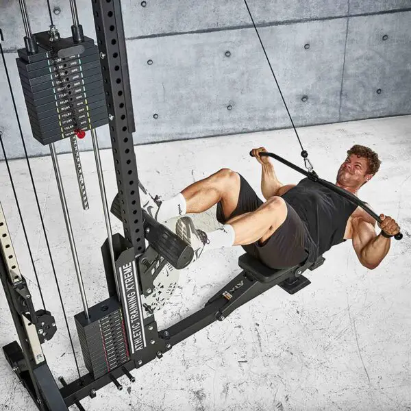 A man in athletic gear is using an ATX® Lat Bar Blackline on a rowing machine against a textured concrete wall, creating an industrial look. The machine includes weights and cables for a full workout.
