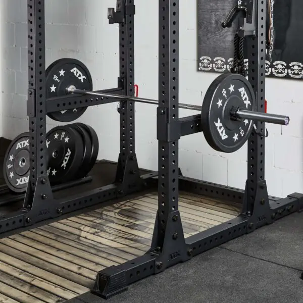 A weightlifting rack with a loaded barbell featuring ATX branding on the plates. The setup includes ATX® Spotter Bars 75cm on a wooden platform, with extra plates stacked on the left. The area is surrounded by gym equipment and chain decor on the wall.