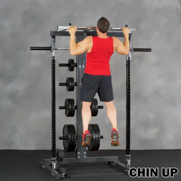 A person in a red tank top and black shorts performs a chin-up on the Ironmaster IM2000 Self-Spotting Smith Machine, with various weight plates visible. The floor is covered with black rubber matting. Text reads CHIN UP.