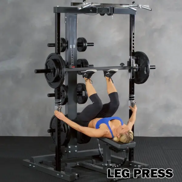 Clad in athletic wear, a person uses the Ironmaster IM2000 Self-Spotting Smith Machine to perform a vertical leg press, pushing a weighted platform with their feet while lying on a bench. Various weight plates surround them with LEG PRESS displayed at the bottom.