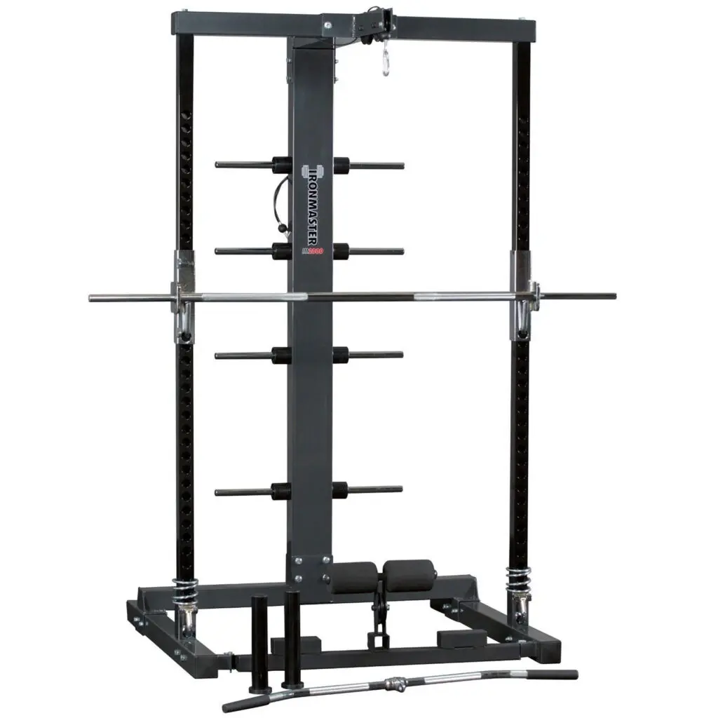 The Ironmaster IM2000 Self-Spotting Smith Machine features adjustable safety bars, a pull-up bar, and multiple weight plate holders. A barbell rests on the safety hooks, with attachments like a curl bar and handle lying before it.