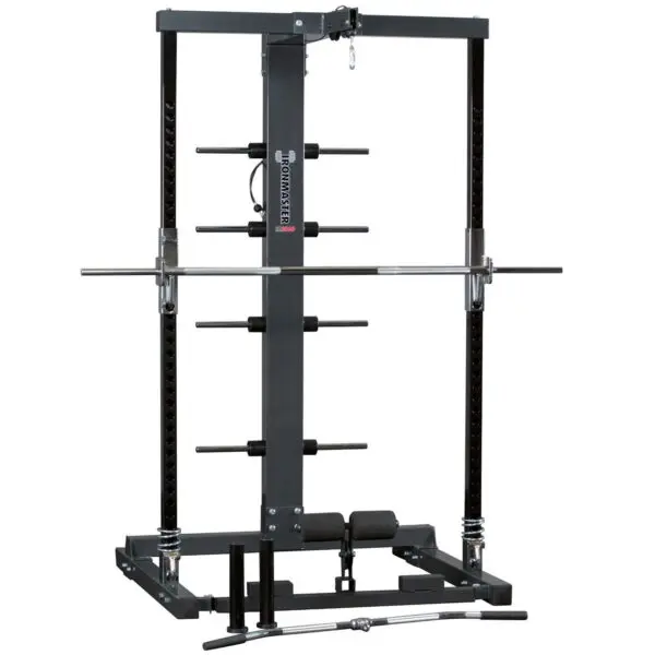 The Ironmaster IM2000 Self-Spotting Smith Machine features adjustable safety bars, a pull-up bar, and multiple weight plate holders. A barbell rests on the safety hooks, with attachments like a curl bar and handle lying before it.