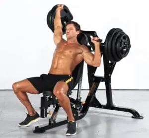 A muscular man is seated on a machine in the gym, performing a shoulder press with weight plates. He is shirtless, wearing black shorts with yellow stripes, and black sneakers. The background is plain white.