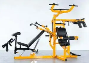 A multi-station home gym with a yellow frame and black cushioning against a plain background. It features an adjustable bench, pulleys, and various attachments for strength training exercises.