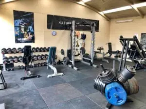 A spacious gym with weightlifting equipment. A rack of dumbbells is on the left, with benches and a squat rack in the center. Weight plates and a leg press machine are visible. Motivational posters hang on the walls.