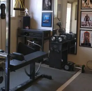 A home gym with various equipment, including a bench press, weight machine, and seated row machine. Walls are decorated with framed posters and a vintage Macintosh computer is placed on a shelf. A mannequin stands in a corner.