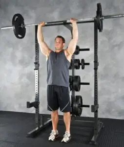 A man in a sleeveless shirt and shorts performs an overhead press with a barbell in a gym. He stands inside a power rack with weight plates on racks behind him, on a rubber mat floor.