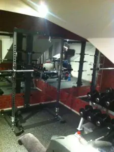 A home gym with a squat rack, dumbbells, and mirrored walls. Gym equipment is neatly arranged, including weights on a rack. A towel and water bottle are visible on a treadmill in the foreground.