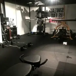 A dimly lit gym with weightlifting equipment, including benches, barbells, and a squat rack. A motivational quote on the wall reads, Sweat is fat crying. The floor is covered with black rubber mats.