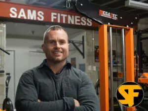 A person standing in a gym with arms crossed, smiling. Theres a weight machine in the background and a sign reading SAMS FITNESS. A logo with the letters SF is visible in the corner.