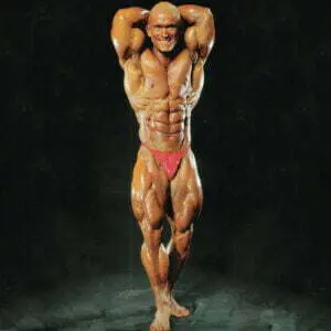 A bodybuilder posing on a dark background, wearing red trunks. The individual has a muscular build, showcasing defined abs and biceps, with hands clasped behind the head and legs slightly apart, highlighting their effort to build massive legs.