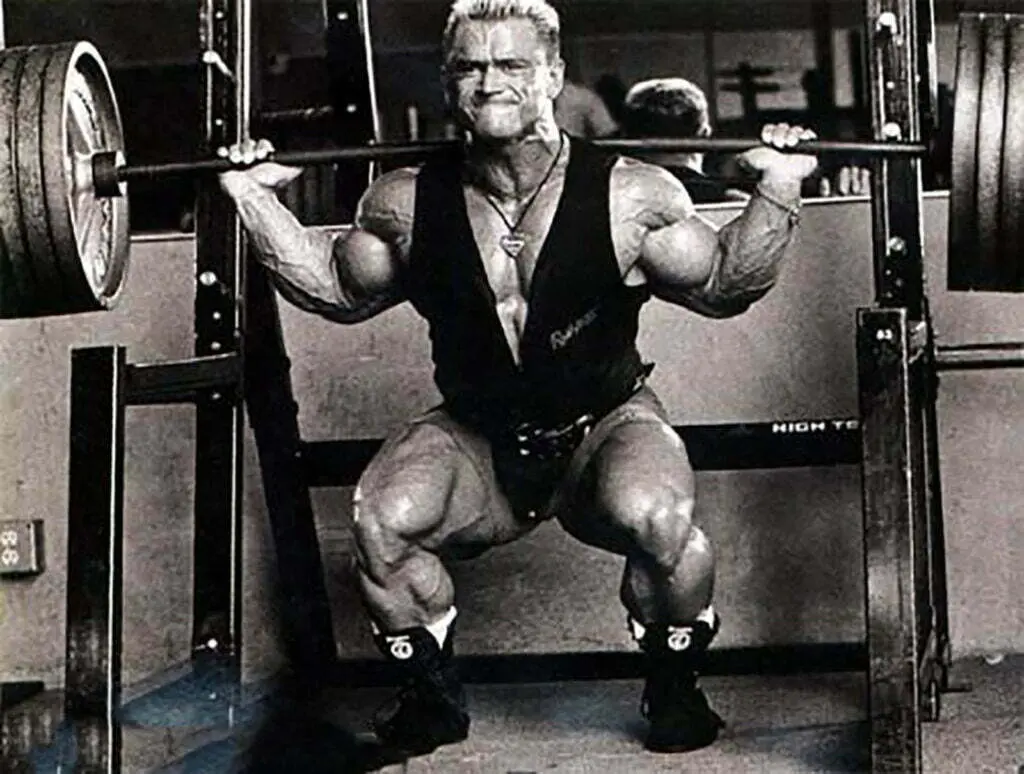 A muscular individual performs a heavy barbell squat in a gym, determined to build massive legs. Dressed in a sleeveless top, shorts, and a weightlifting belt, their intense expression is focused on the lift. Fitness equipment fills the background.