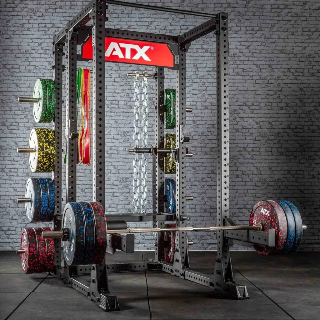 A gym setup featuring a power rack with multiple weight plates and chains. The equipment is positioned against a brick wall, with a red ATX logo above the rack. The weights have a colorful speckled design.