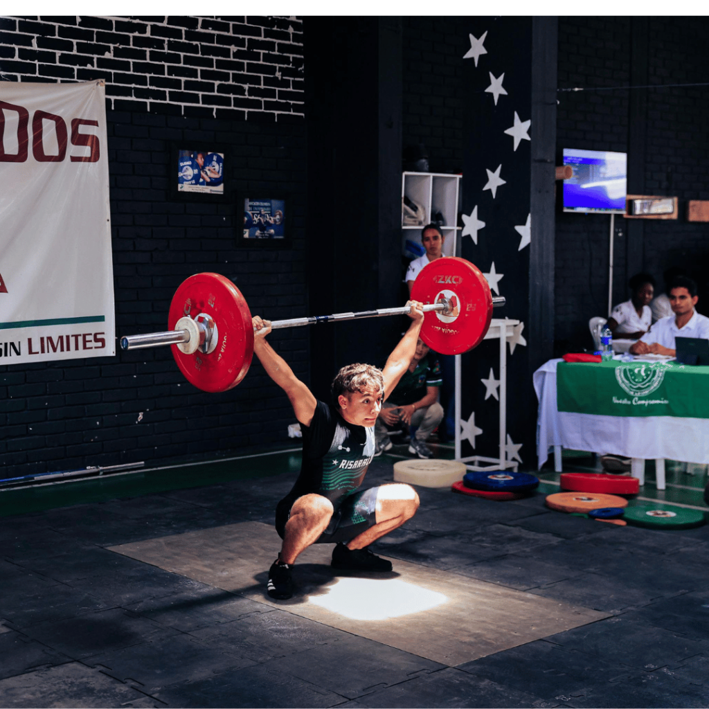 A male weightlifter in a black outfit performs a snatch, squatting low while lifting a barbell with red weights overhead in an indoor gym. Judges and weights are visible in the background.