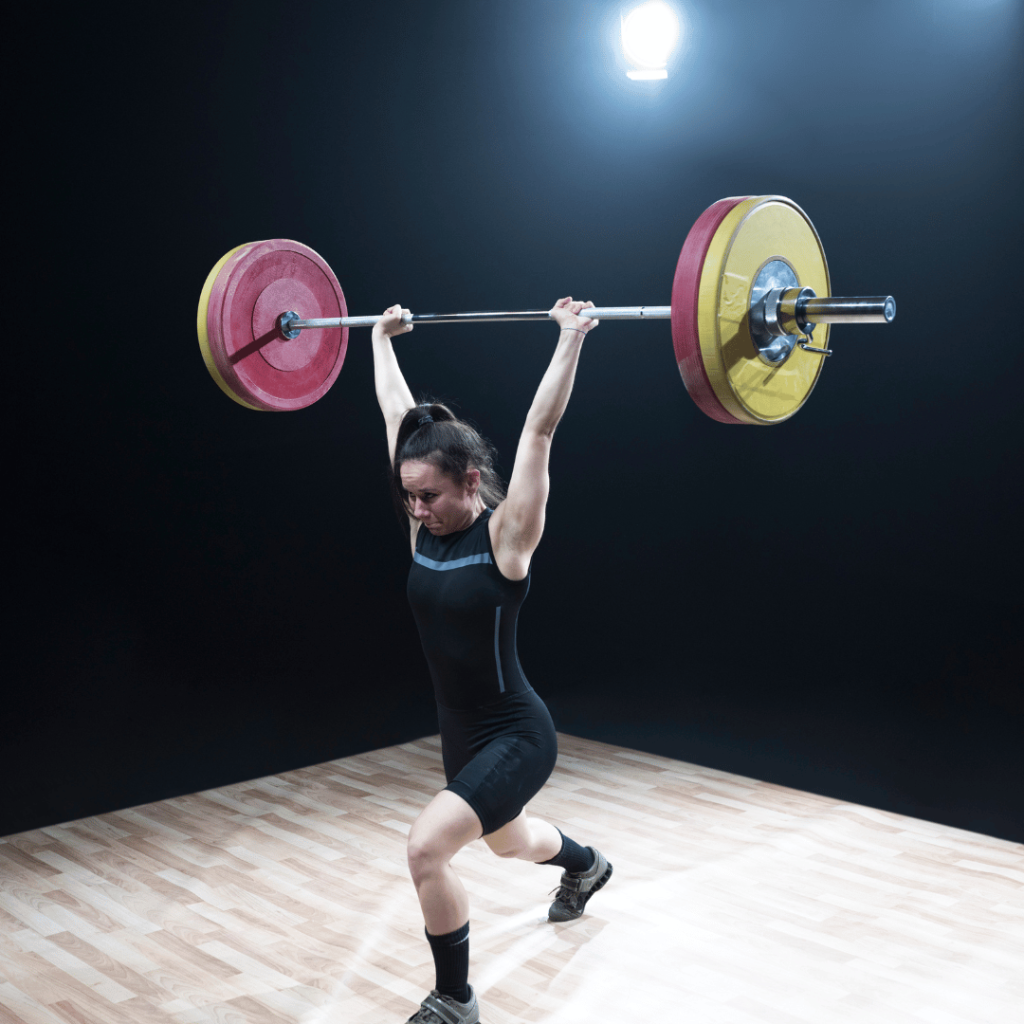 A woman in athletic gear performs a split jerk, lifting a loaded barbell overhead on a wooden floor under bright lights, with a focused expression and strong stance.