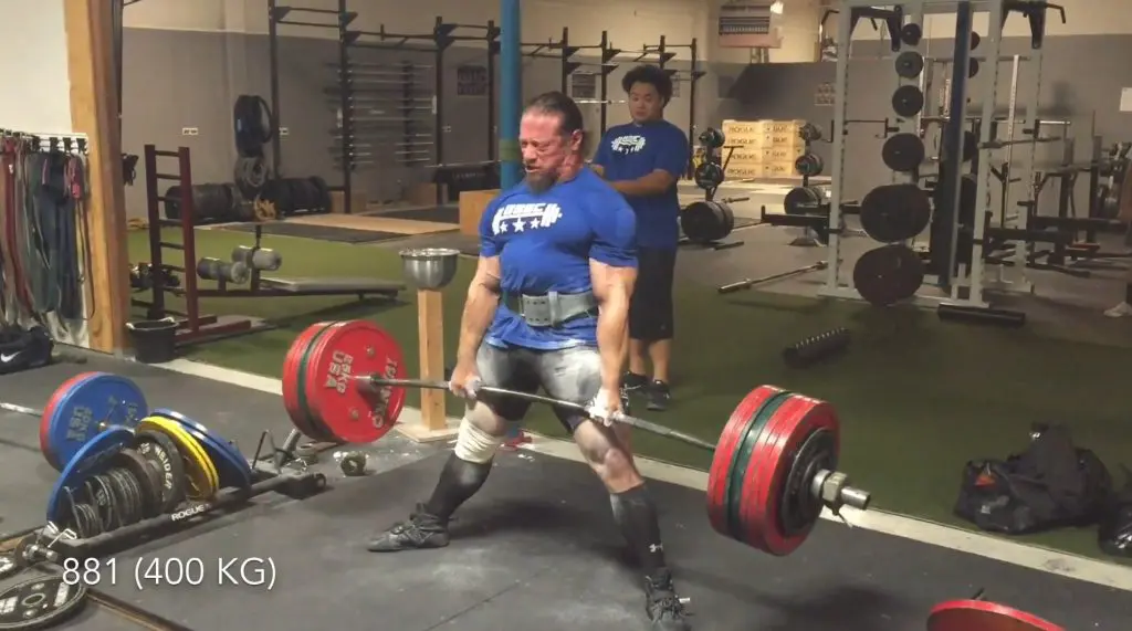 A man wearing a blue shirt and lifting belt is performing a heavy deadlift with a loaded barbell in a gym. Another person stands behind him observing. Text on the image reads 881 (400 KG).