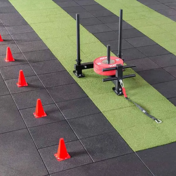 A gym sled with red weights and a harness rests on a green-and-black GymFloor RTS 50x50@20mm System. Red cones are aligned beside the sled, indicating a training path.