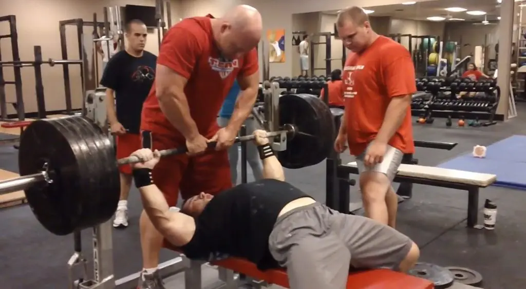 A man lies on a bench press lifting a heavily loaded barbell, assisted by a spotter. Three other men stand nearby in a gym, watching and ready to help. The gym has weights and equipment in the background.