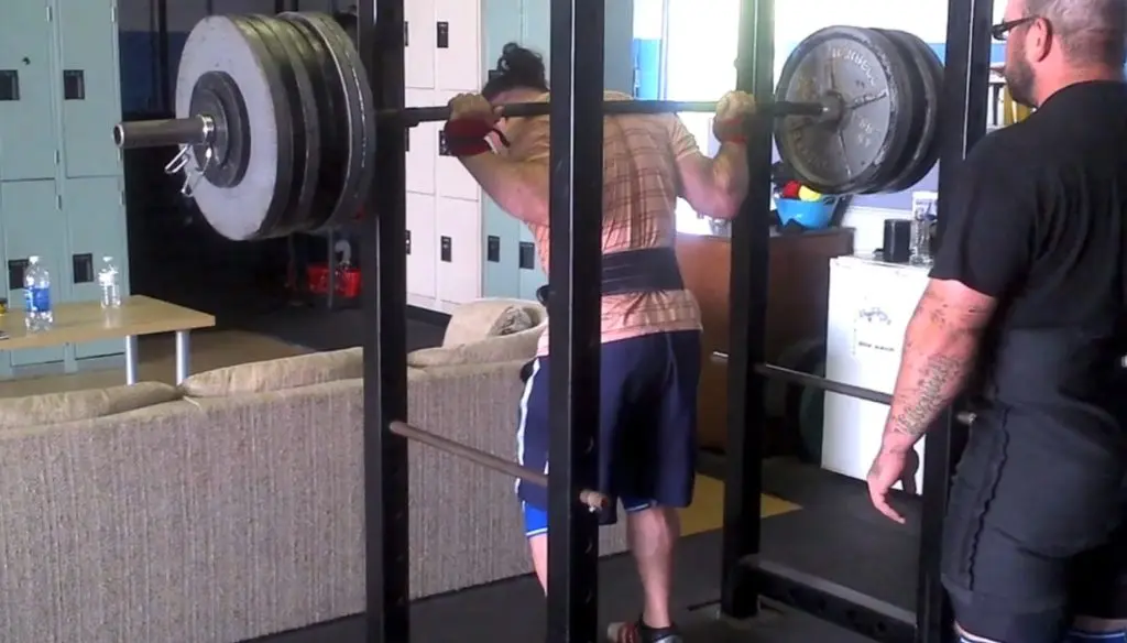 A person wearing gym clothes and a lifting belt prepares to squat a heavily loaded barbell on a squat rack, while another person stands nearby spotting them in a gym setting.