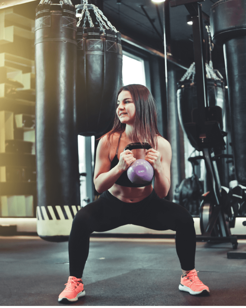 A woman in athletic wear performs a squat while holding a purple kettlebell in a gym with punching bags and gym equipment in the background.