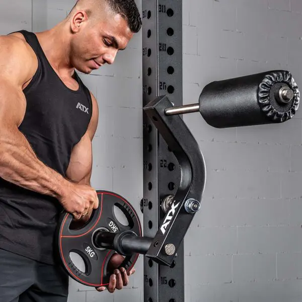 A person in a black tank top adjusts the ATX® Neck Trainer Add On on a barbell attached to a wall-mounted fitness machine, focusing intently against the backdrop of a light gray brick wall.