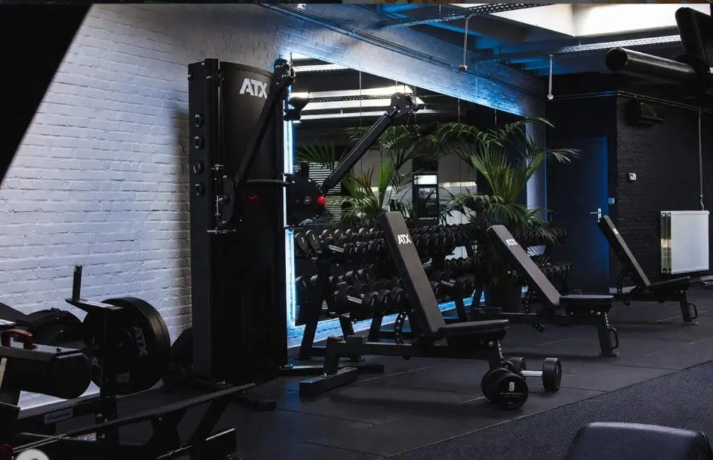 A modern gym features the ATX® Flat Incline Bench 610, dumbbells, and a cable machine against a white brick wall. Large mirrors, potted plants, and black flooring create a clean, organized workout space.