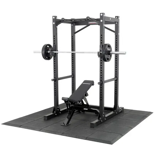 The Shock & Sound Tile 30mm System supports a power rack loaded with a barbell and weights on its black rubber mat, with an adjustable bench inside the rack for versatile strength training exercises.