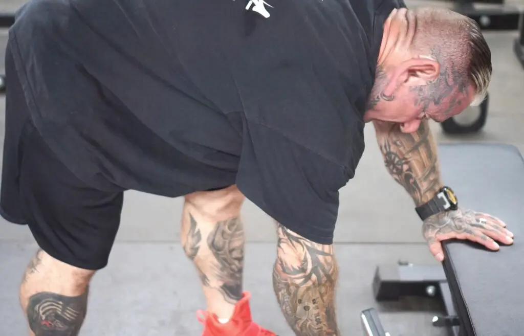 A person with tattoos on their arms and legs is leaning over a bench in a gym. They are wearing a black t-shirt, black shorts, and red sneakers. Their hand is resting on the bench, and they are focused on a workout.