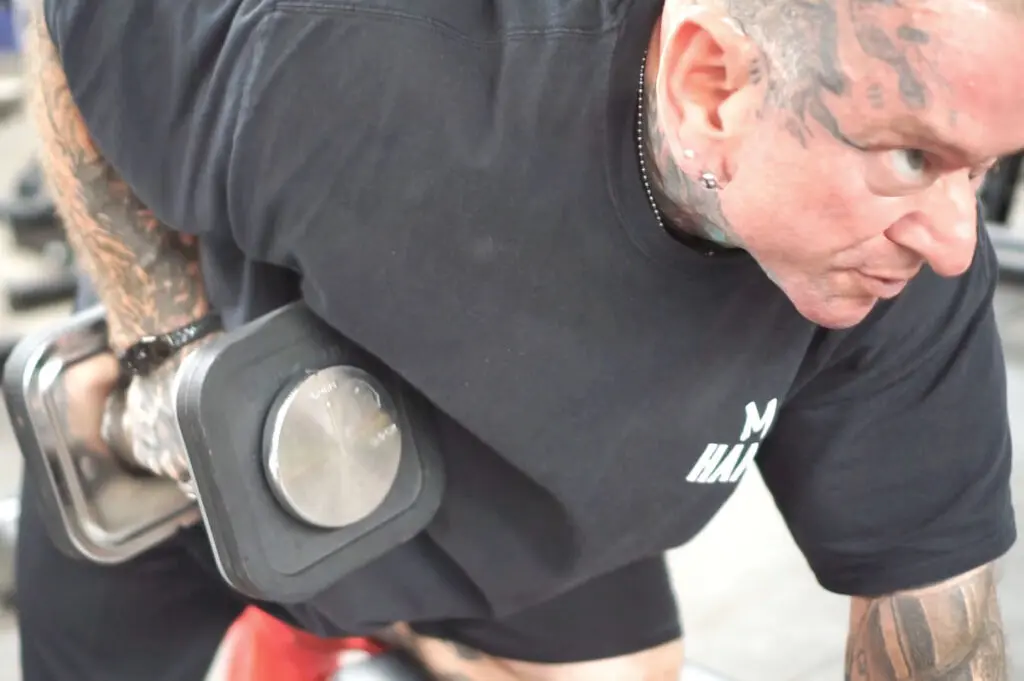 A muscular person with tattoos is performing a dumbbell row exercise in the gym. They are wearing a black shirt and focusing intently on the movement.