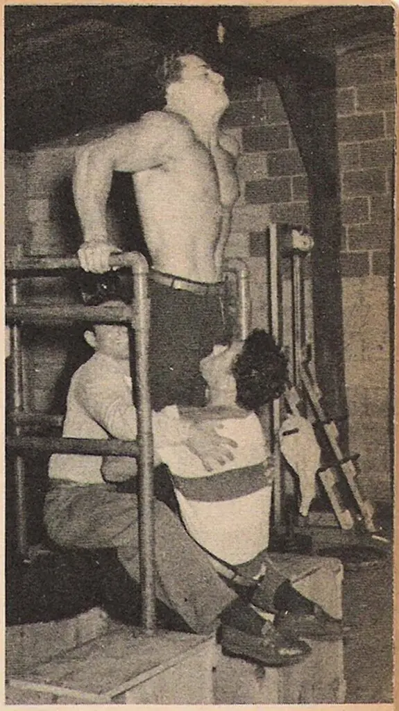 A shirtless man performs an exercise using parallel bars, while two men help by holding his legs for support. The setting appears to be a gym or workout area with brick walls and equipment in the background.
