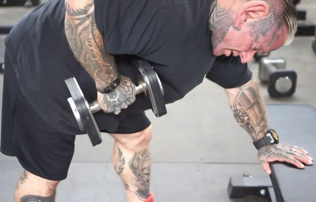 A tattooed person with short hair, wearing a black shirt and shorts, performs a bent-over dumbbell row at the gym. Their left hand grips the weight while the right hand is placed on a bench for support.